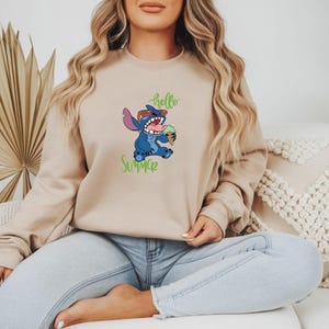 Puede incluir: Sudadera beige con un personaje de dibujos animados azul con gafas de sol y un helado. Las palabras "hello summer" est&aacute;n escritas en verde. La sudadera es de color neutro y tiene mangas largas.