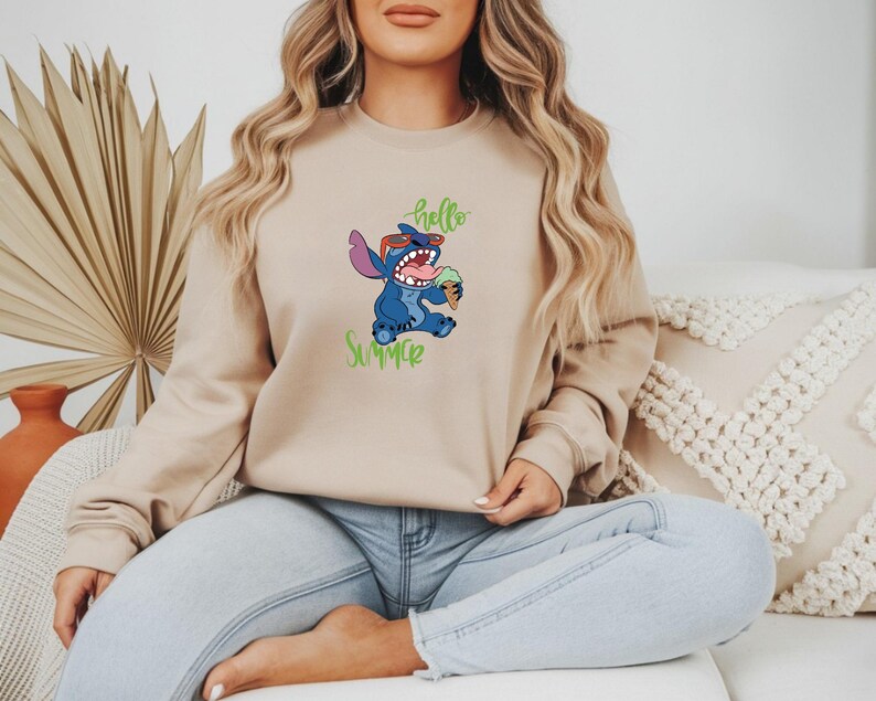 Puede incluir: Sudadera beige con un personaje de dibujos animados azul con gafas de sol y un helado. Las palabras "hello summer" est&aacute;n escritas en verde. La sudadera es de color neutro y tiene mangas largas.