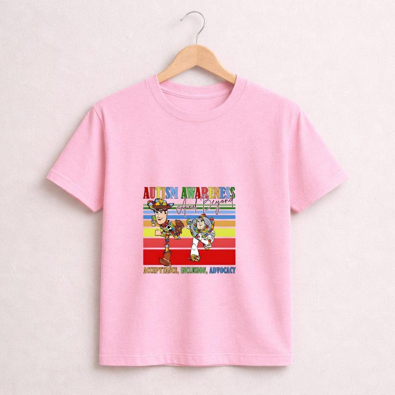 May include: A light pink t-shirt featuring a colorful graphic with the text "AUTISM AWARENESS And Beyond". The graphic includes cartoon characters and the words "ACCEPTANCE, INCLUSION, ADVOCACY". The shirt is hanging on a wooden hanger.