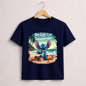Pu&ograve; includere: T-shirt blu navy con un personaggio dei cartoni animati su una tavola da surf sulla spiaggia. Il personaggio &egrave; blu con grandi orecchie e sorride. Il testo "Too Cute to be this Troublemaker" &egrave; sopra il personaggio.