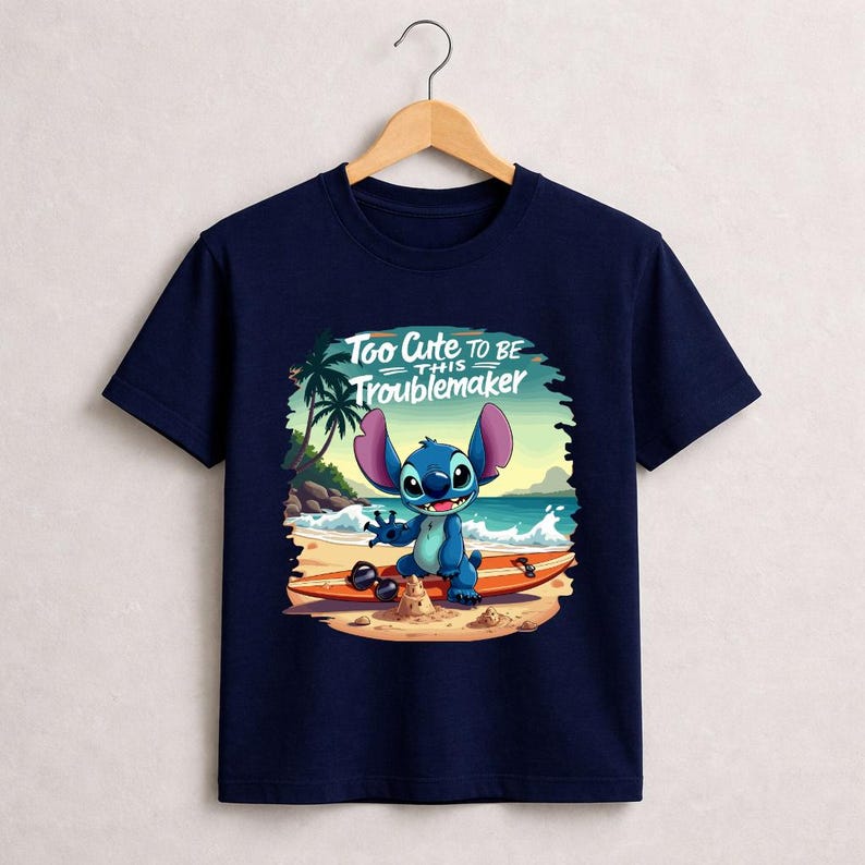 Pu&ograve; includere: T-shirt blu navy con un personaggio dei cartoni animati su una tavola da surf sulla spiaggia. Il personaggio &egrave; blu con grandi orecchie e sorride. Il testo "Too Cute to be this Troublemaker" &egrave; sopra il personaggio.