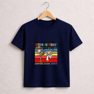 May include: Navy blue t-shirt featuring a colorful graphic with the text "AUTISM AWARENESS" and the words "ACCEPTANCE, INCLUSION, ADVOCACY". The graphic includes cartoon characters. The shirt is hanging on a wooden hanger.