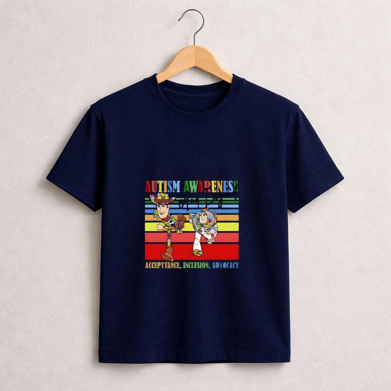 May include: Navy blue t-shirt featuring a colorful graphic with the text "AUTISM AWARENESS" and the words "ACCEPTANCE, INCLUSION, ADVOCACY". The graphic includes cartoon characters. The shirt is hanging on a wooden hanger.