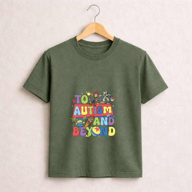Op de afbeelding: Olijfgroen T-shirt met een kleurrijke afbeelding met de tekst "TO AUTISM AND BEYOND" met stripfiguren. Het shirt hangt aan een houten hanger tegen een effen achtergrond.
