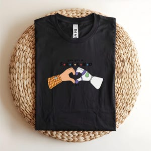 May include: Black t-shirt featuring a graphic of two cartoon hands forming a heart. One hand has a plaid sleeve, the other is a robotic arm. The text reads "you got a friend in me."