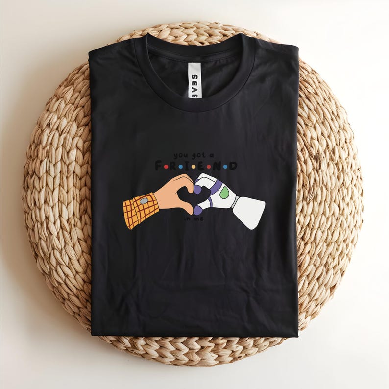 May include: Black t-shirt featuring a graphic of two cartoon hands forming a heart. One hand has a plaid sleeve, the other is a robotic arm. The text reads "you got a friend in me."