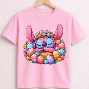 May include: Pink t-shirt featuring a cartoon character with large ears, blue skin, and a floral crown, nestled among colorful Easter eggs. The design is centered on the shirt, creating a festive and playful look.
