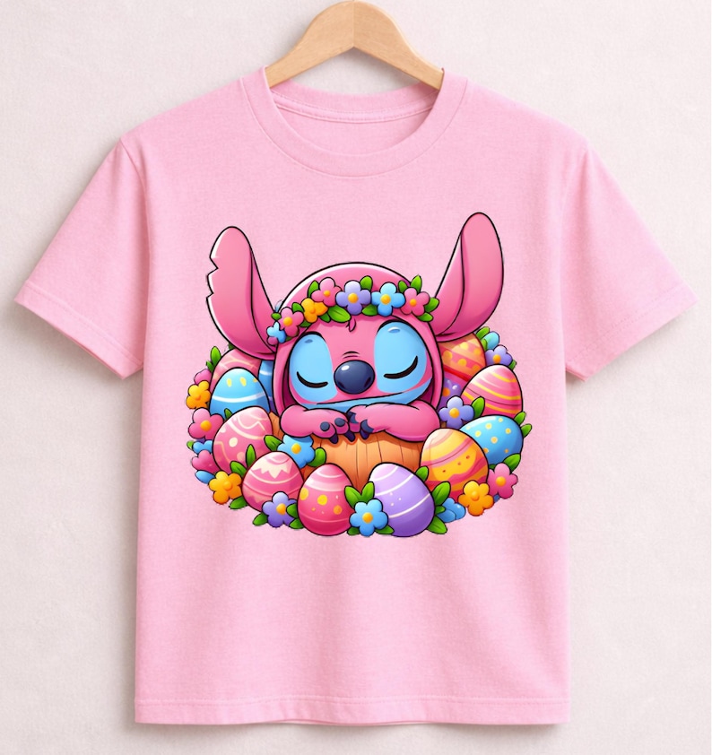 May include: Pink t-shirt featuring a cartoon character with large ears, blue skin, and a floral crown, nestled among colorful Easter eggs. The design is centered on the shirt, creating a festive and playful look.