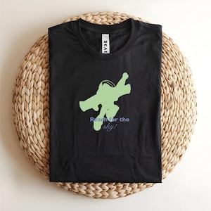 May include: Black t-shirt featuring a green graphic of a figure in a flying pose and the text "Reach for the sky!" in blue. The shirt is folded on a woven, circular surface.