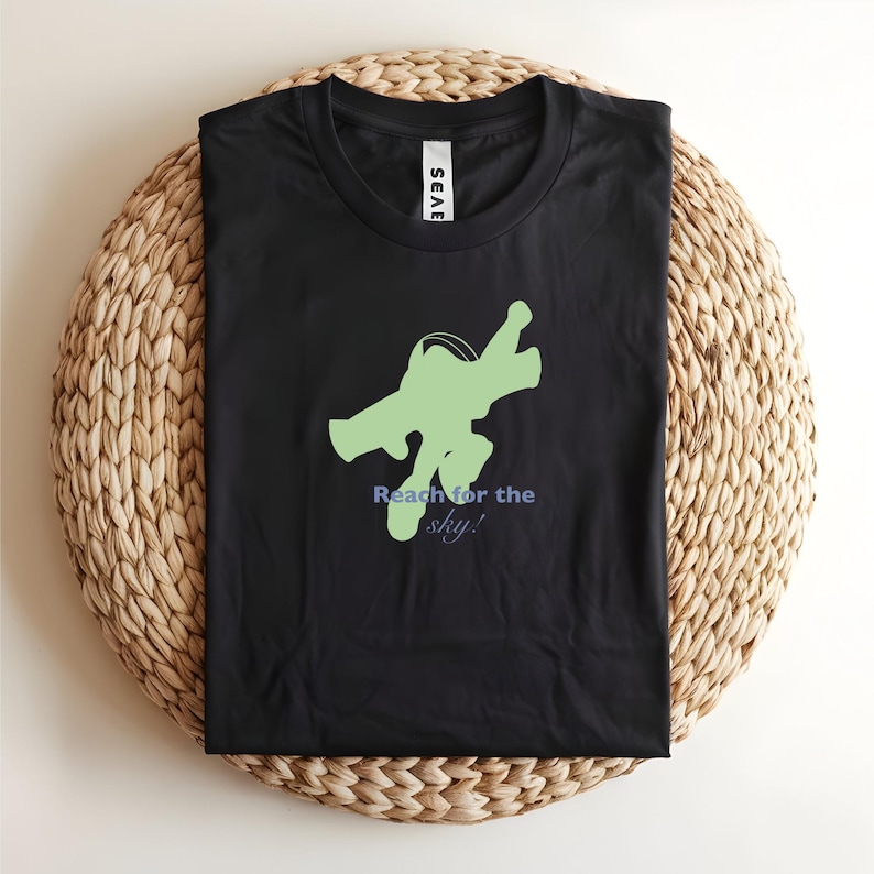 May include: Black t-shirt featuring a green graphic of a figure in a flying pose and the text "Reach for the sky!" in blue. The shirt is folded on a woven, circular surface.