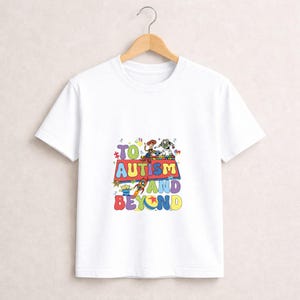 Op de afbeelding: Wit T-shirt met een kleurrijke afbeelding met personages en de tekst "TO AUTISM AND BEYOND". Het ontwerp bevat een raket, sterren en een retro lettertype. Het shirt hangt aan een houten hanger.
