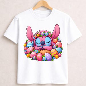 May include: White t-shirt featuring a cartoon character with pink fur, blue face, and a flower crown, nestled among colorful Easter eggs. The design is vibrant and festive, perfect for Easter celebrations.