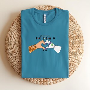 May include: Teal t-shirt with a graphic of two cartoon hands forming a heart shape. One hand is human, the other robotic. The text "you got a FRIEND in me" is above and below the hands.