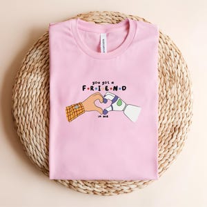 May include: A light pink t-shirt featuring the text "you got a FRIEND in me" with a heart made of two cartoon hands. One hand has a plaid sleeve, the other is a robot hand. The shirt is folded on a woven mat.