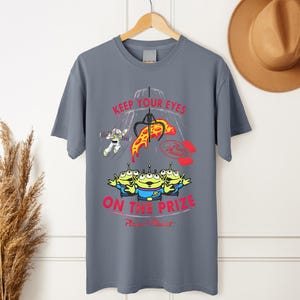 May include: Gray t-shirt featuring a graphic from the movie Toy Story. The design includes the text "KEEP YOUR EYES ON THE PRIZE" with images of Buzz Lightyear, the Little Green Men, and a pizza slice.