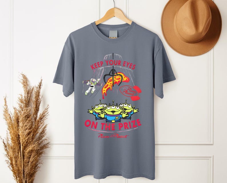 May include: Gray t-shirt featuring a graphic from the movie Toy Story. The design includes the text "KEEP YOUR EYES ON THE PRIZE" with images of Buzz Lightyear, the Little Green Men, and a pizza slice.