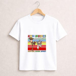 May include: White t-shirt featuring a colorful graphic with the text "Autism Awareness and Beyond." The design includes cartoon characters and the words "Acceptance, Inclusion, Advocacy" on a striped background.