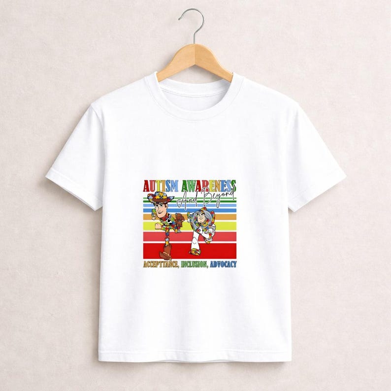 May include: White t-shirt featuring a colorful graphic with the text "Autism Awareness and Beyond." The design includes cartoon characters and the words "Acceptance, Inclusion, Advocacy" on a striped background.