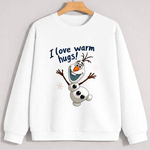 May include: White sweatshirt featuring a cartoon image of Olaf from Frozen. The image includes the text "I love warm hugs!" in blue. Olaf is depicted with a smiling face, twig arms, and a carrot nose. The sweatshirt has a crew neck and long sleeves.