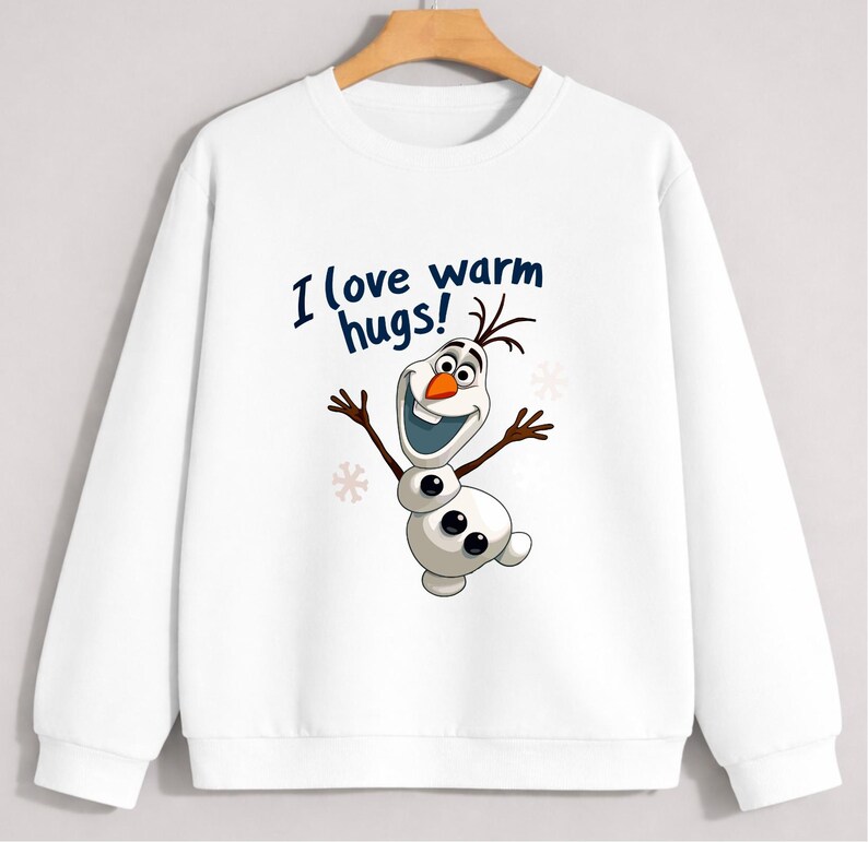 May include: White sweatshirt featuring a cartoon image of Olaf from Frozen. The image includes the text "I love warm hugs!" in blue. Olaf is depicted with a smiling face, twig arms, and a carrot nose. The sweatshirt has a crew neck and long sleeves.