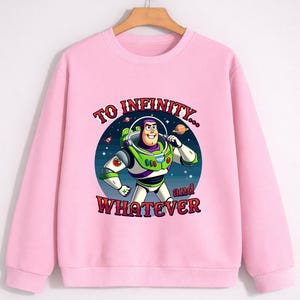 Op de afbeelding: Een lichtroze sweatshirt met een afbeelding van Buzz Lightyear uit Toy Story. De cartoonafbeelding staat tegen een sterrenachtergrond en bevat de tekst "TO INFINITY... and WHATEVER". De sweatshirt heeft een ronde hals en lange mouwen.