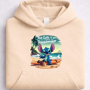 Puede incluir: Sudadera con capucha beige con un gr&aacute;fico de un personaje de dibujos animados azul en una tabla de surf en la playa. El gr&aacute;fico incluye el texto "Too Cute To Be This Troublemaker". La sudadera tiene un bolsillo delantero y una capucha.