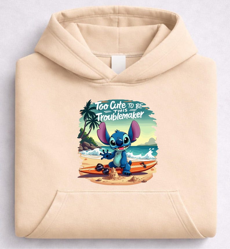 Puede incluir: Sudadera con capucha beige con un gr&aacute;fico de un personaje de dibujos animados azul en una tabla de surf en la playa. El gr&aacute;fico incluye el texto "Too Cute To Be This Troublemaker". La sudadera tiene un bolsillo delantero y una capucha.