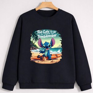 Puede incluir: Sudadera negra con un personaje de dibujos animados en una tabla de surf en la playa. El personaje azul tiene orejas grandes y est&aacute; sonriendo. El texto "Too Cute to be this Troublemaker" est&aacute; encima del personaje.
