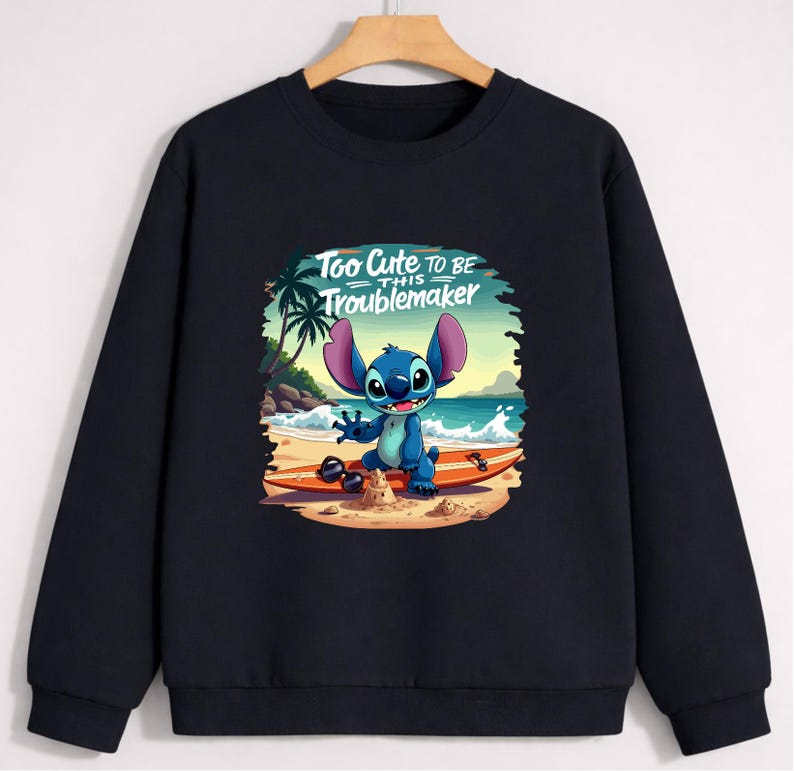 Puede incluir: Sudadera negra con un personaje de dibujos animados en una tabla de surf en la playa. El personaje azul tiene orejas grandes y est&aacute; sonriendo. El texto "Too Cute to be this Troublemaker" est&aacute; encima del personaje.