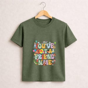 May include: Olive green t-shirt with the phrase "YOU'VE GOT A FRIEND IN ME" in colorful, playful lettering. The design features cartoon characters and balloons, creating a cheerful and nostalgic vibe.