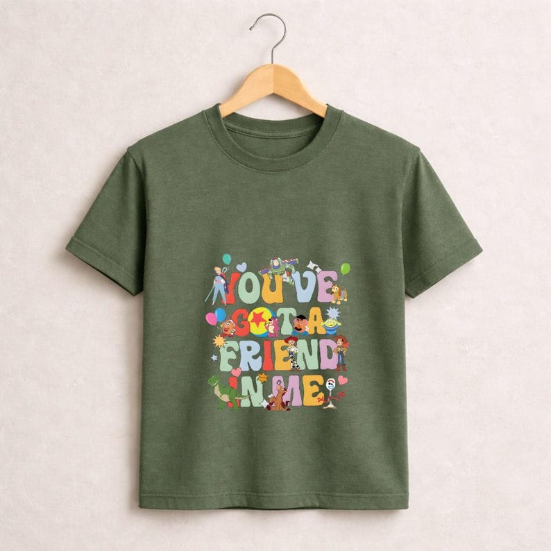 May include: Olive green t-shirt with the phrase "YOU'VE GOT A FRIEND IN ME" in colorful, playful lettering. The design features cartoon characters and balloons, creating a cheerful and nostalgic vibe.