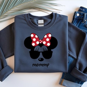 Może przedstawiać: Granatowa bluza z nadrukiem Myszki Minnie. Wz&oacute;r zawiera czerwoną kokardę w białe grochy, czarne okulary przeciwsłoneczne i napis "mamusia". Bluza jest połączona z niebieskimi dżinsami.