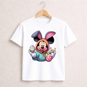 May include: White t-shirt featuring a cartoon Mickey Mouse character dressed as an Easter bunny. Mickey is wearing a hooded sweatshirt and holding colorful Easter eggs and flowers. The design is vibrant and playful, perfect for Easter.