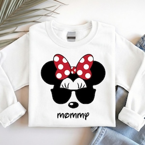 Może przedstawiać: Biała bluza z długim rękawem z motywem Myszki Minnie. Projekt zawiera czarną sylwetkę głowy Myszki Minnie, okulary przeciwsłoneczne i czerwoną kokardę w białe grochy. Słowo "mommy" jest wydrukowane poniżej.