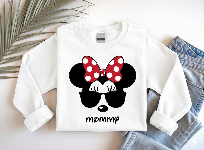 Może przedstawiać: Biała bluza z długim rękawem z motywem Myszki Minnie. Projekt zawiera czarną sylwetkę głowy Myszki Minnie, okulary przeciwsłoneczne i czerwoną kokardę w białe grochy. Słowo "mommy" jest wydrukowane poniżej.