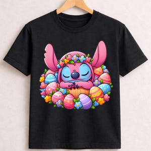 May include: Black t-shirt featuring a cartoon character with pink ears and blue skin, surrounded by colorful Easter eggs and flowers. The character is wearing a flower crown and is nestled in a basket. A fun, festive design for Easter.