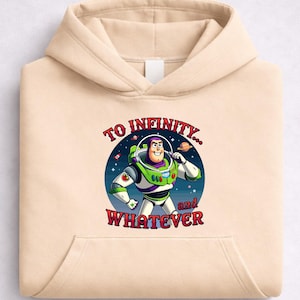 Op de afbeelding: Beige hoodie met een afbeelding van Buzz Lightyear en de tekst "TO INFINITY... and WHATEVER". Het ontwerp toont een ruimteachtergrond en het personage in een groen en wit ruimtepak.