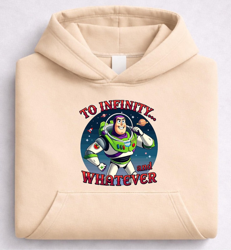 Op de afbeelding: Beige hoodie met een afbeelding van Buzz Lightyear en de tekst "TO INFINITY... and WHATEVER". Het ontwerp toont een ruimteachtergrond en het personage in een groen en wit ruimtepak.