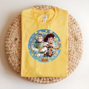 Puede incluir: Camiseta amarilla con un gr&aacute;fico circular de Buzz Lightyear y Woody de Toy Story. El gr&aacute;fico est&aacute; enmarcado por un borde azul con estrellas y las palabras "Toy Story". La camiseta est&aacute; hecha de un material suave.