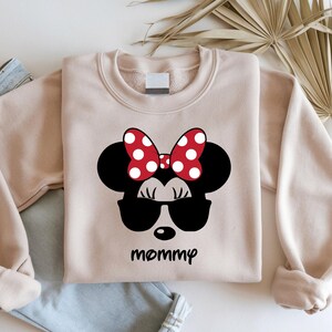 Może przedstawiać: Jasnobrązowa bluza z czarnym motywem Myszki Minnie w okularach przeciwsłonecznych i czerwoną kokardą w białe grochy. Słowo "mommy" jest wydrukowane poniżej na czarno.