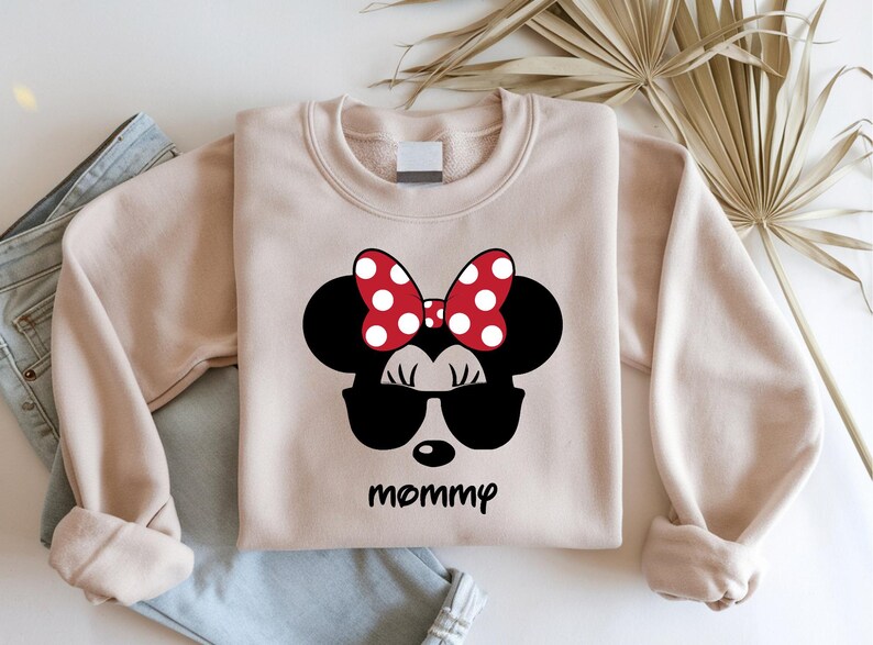 Może przedstawiać: Jasnobrązowa bluza z czarnym motywem Myszki Minnie w okularach przeciwsłonecznych i czerwoną kokardą w białe grochy. Słowo "mommy" jest wydrukowane poniżej na czarno.
