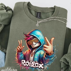 Puede incluir: Sudadera verde militar con un gr&aacute;fico colorido de un personaje de Roblox con gafas de sol y un signo de la paz. La palabra "ROBLOX" se muestra en blanco. Un collar plateado con un colgante est&aacute; sobre la sudadera.