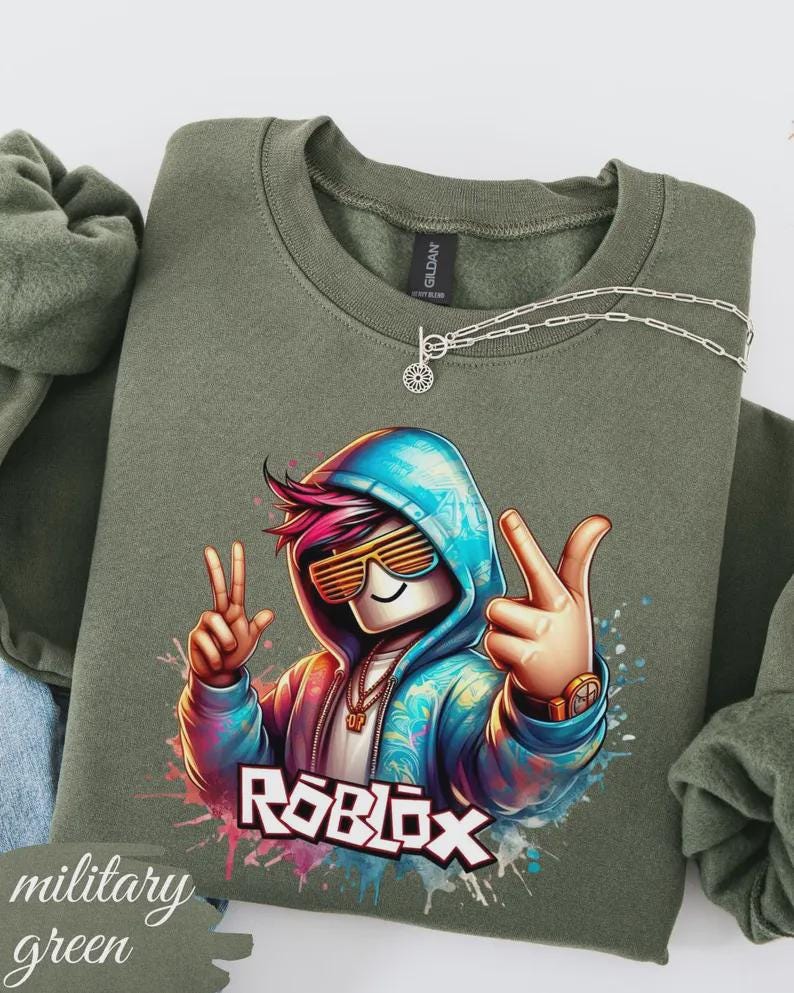 Puede incluir: Sudadera verde militar con un gr&aacute;fico colorido de un personaje de Roblox con gafas de sol y un signo de la paz. La palabra "ROBLOX" se muestra en blanco. Un collar plateado con un colgante est&aacute; sobre la sudadera.