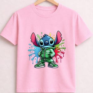 May include: A light pink t-shirt featuring a cartoon character in a green tracksuit with a colorful paint splatter design. The character is blue with large ears and is wearing green sneakers.