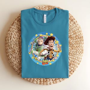 Puede incluir: Camiseta de color turquesa con un gr&aacute;fico circular de Buzz Lightyear y Woody de Toy Story. El gr&aacute;fico incluye el texto "TOY STORY" en un dise&ntilde;o colorido. La camiseta est&aacute; hecha de un material suave y se muestra sobre una superficie tejida.