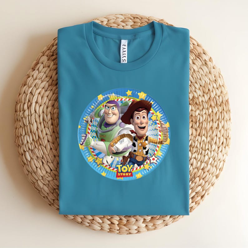 Puede incluir: Camiseta de color turquesa con un gr&aacute;fico circular de Buzz Lightyear y Woody de Toy Story. El gr&aacute;fico incluye el texto "TOY STORY" en un dise&ntilde;o colorido. La camiseta est&aacute; hecha de un material suave y se muestra sobre una superficie tejida.