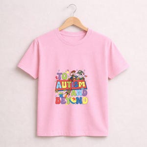 Op de afbeelding: Een lichtroze t-shirt met een kleurrijke afbeelding met de tekst "TO AUTISM AND BEYOND" en Toy Story-personages. Het ontwerp bevat Woody, Buzz Lightyear en andere personages uit de film. Het t-shirt hangt aan een houten hanger.