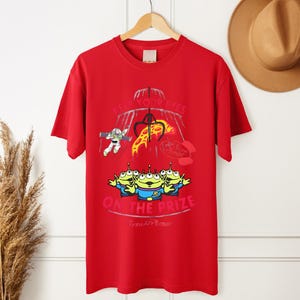 May include: Red t-shirt featuring a graphic from the movie Toy Story. The design includes the text "KEEP YOUR EYES ON THE PRIZE" with images of the Little Green Men, Buzz Lightyear, and a claw machine.
