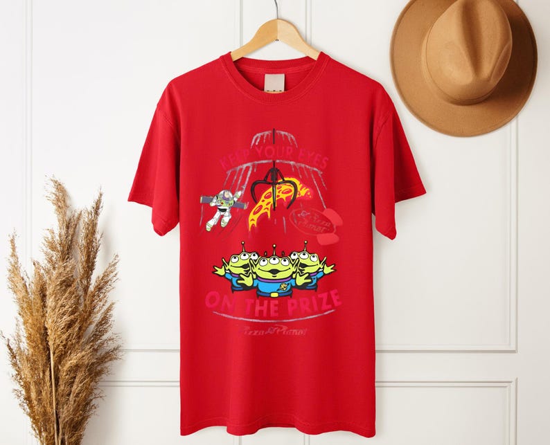 May include: Red t-shirt featuring a graphic from the movie Toy Story. The design includes the text "KEEP YOUR EYES ON THE PRIZE" with images of the Little Green Men, Buzz Lightyear, and a claw machine.