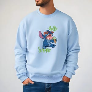 Puede incluir: Sudadera azul claro con un personaje de dibujos animados, Stitch, con gafas de sol y un helado. Las palabras "hello Summer" est&aacute;n escritas en verde arriba y abajo del personaje. La sudadera es de color azul claro liso.